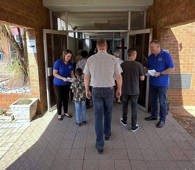 Twee gashere in blou hemde verwelkom mense by die kerk se ingang